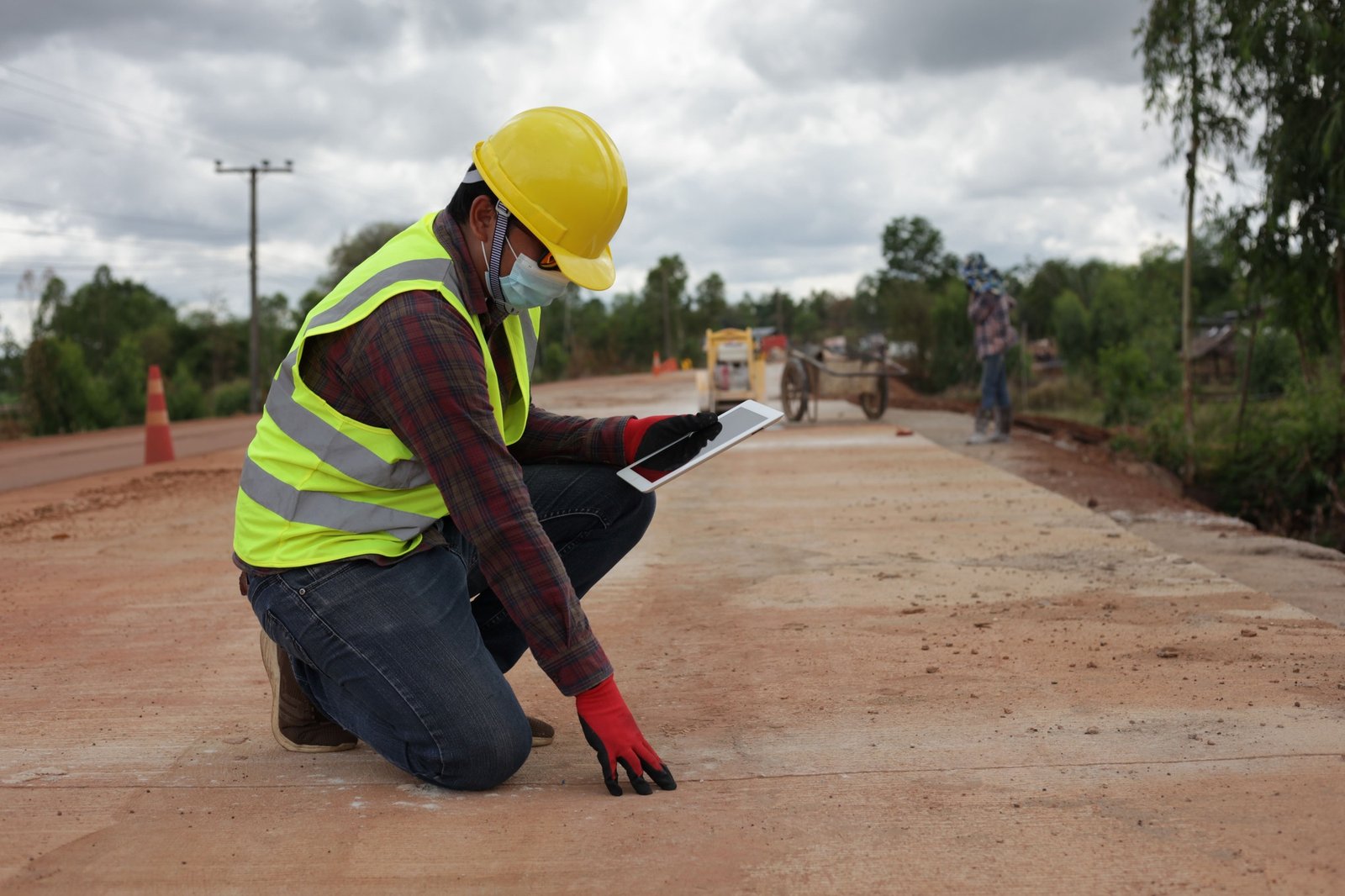A male contractor wearing a helmet with a tablet bends down to inspect a concrete road.